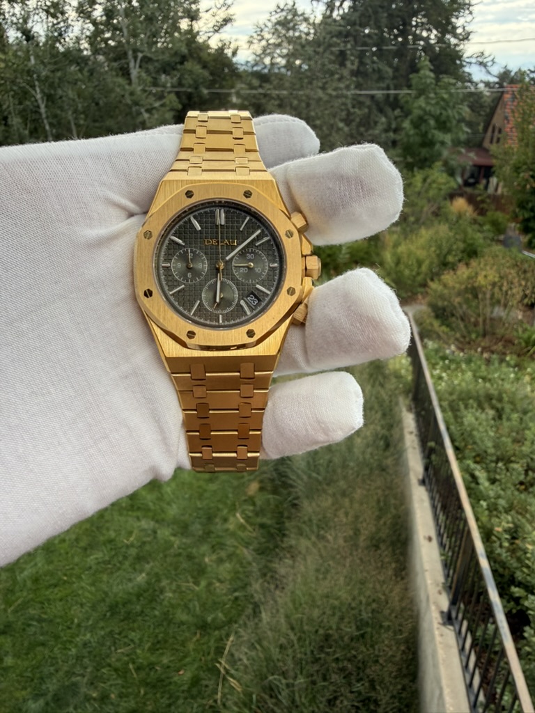 Gold Chronograph Homage — held in a gloved hand with a hillside view behind it