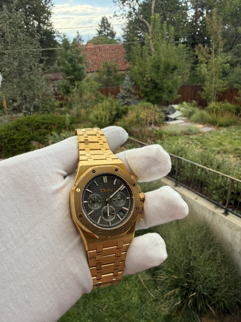 Gold Chronograph Homage — held in a gloved hand overlooking a garden and rooftop
