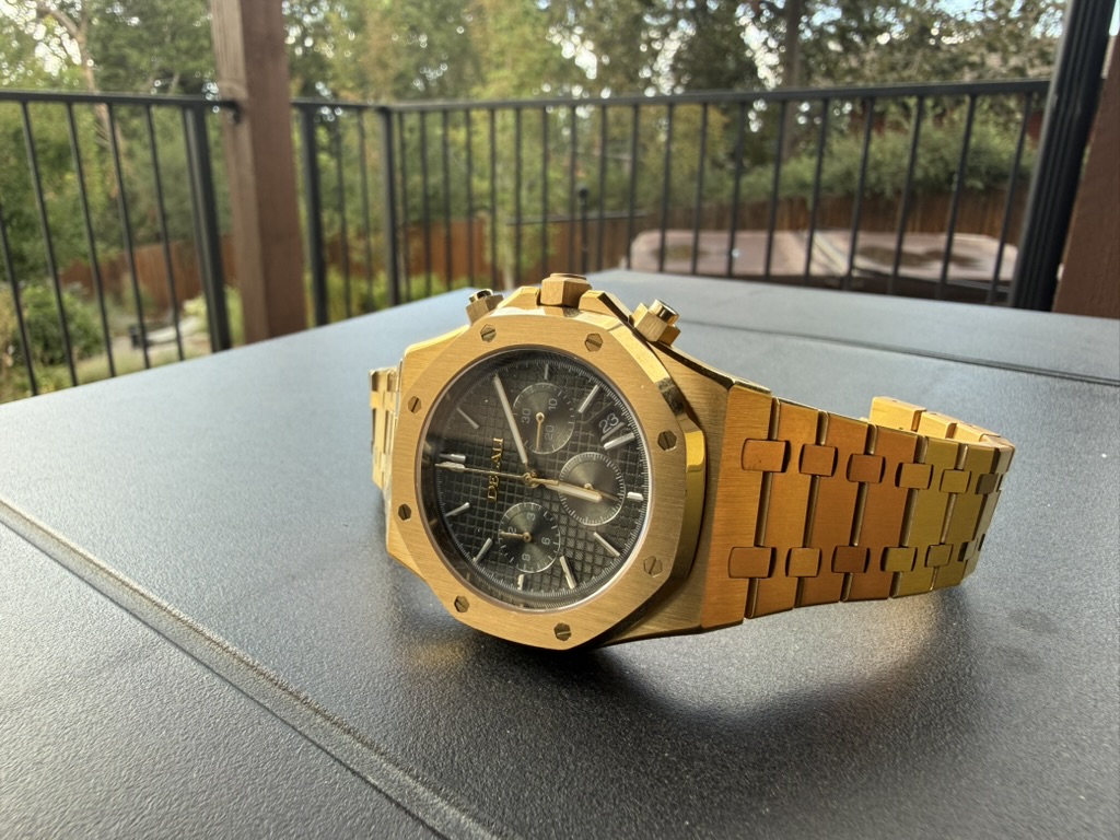 Gold Chronograph Homage — resting on an outdoor table with trees and a deck railing behind it