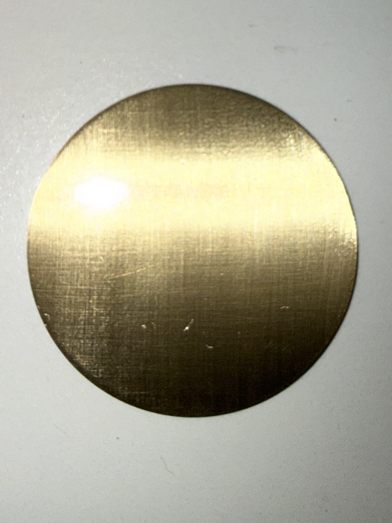 A brass dial blank, before any texture is worked into it.
